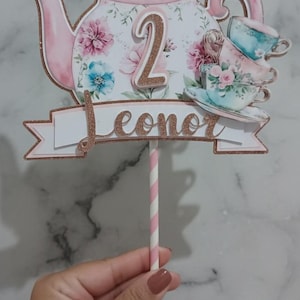 May include: A decorative cake topper featuring a pink teapot with floral designs, a butterfly, and stacked teacups. The number "2" and the name "Leonor" are displayed in glittery gold. A pink and white striped straw supports the topper.
