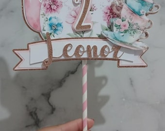 May include: A decorative cake topper featuring a pink teapot with floral designs, a butterfly, and stacked teacups. The number "2" and the name "Leonor" are displayed in glittery gold. A pink and white striped straw supports the topper.
