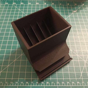 May include: A black plastic dice tower with a rectangular base and a square top. The tower has a series of slots on the inside to guide the dice down.