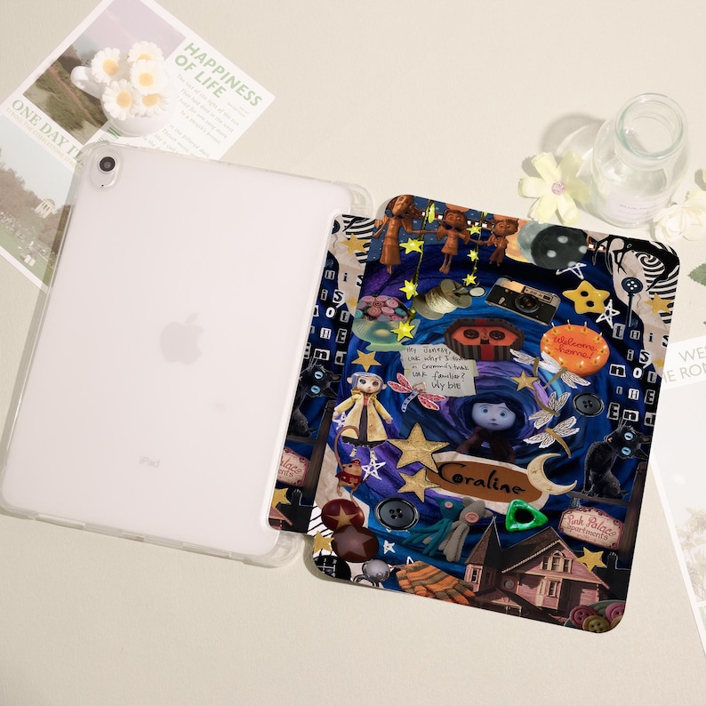 May include: A clear iPad case with a colorful, patterned insert featuring a collage of illustrations and the name "Coraline." The insert has a dark blue background with stars, buttons, and character figures. The iPad is white with the Apple logo.