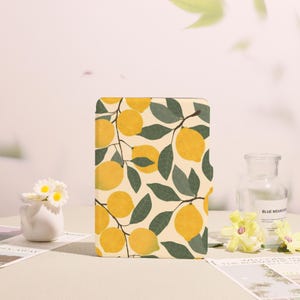 May include: A rectangular, cream-colored case featuring a pattern of yellow lemons and green leaves on branches. The design is repeated across the surface. The case is set against a neutral background with decorative elements.