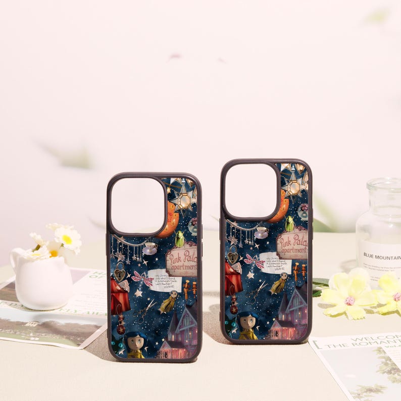 May include: Two dark-rimmed phone cases featuring a dark blue background with a whimsical collage of illustrations including a house, a character with blue hair, a sign reading Pink Palace apartments, stars, scissors, and various small trinkets.