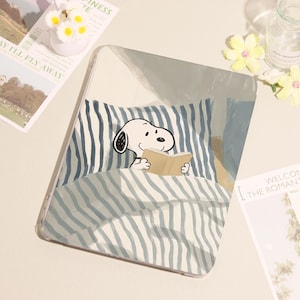 May include: Clear tablet case featuring a cartoon Snoopy character reading a book in bed. The design includes blue and white striped bedding and a light blue pillow. The case has a transparent border.