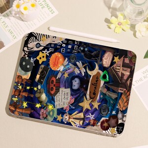 May include: A decorative tablet case featuring a collage design with a dark blue background. The design includes stars, buttons, a camera, and the word "Coraline". A white stylus rests on top of the case.