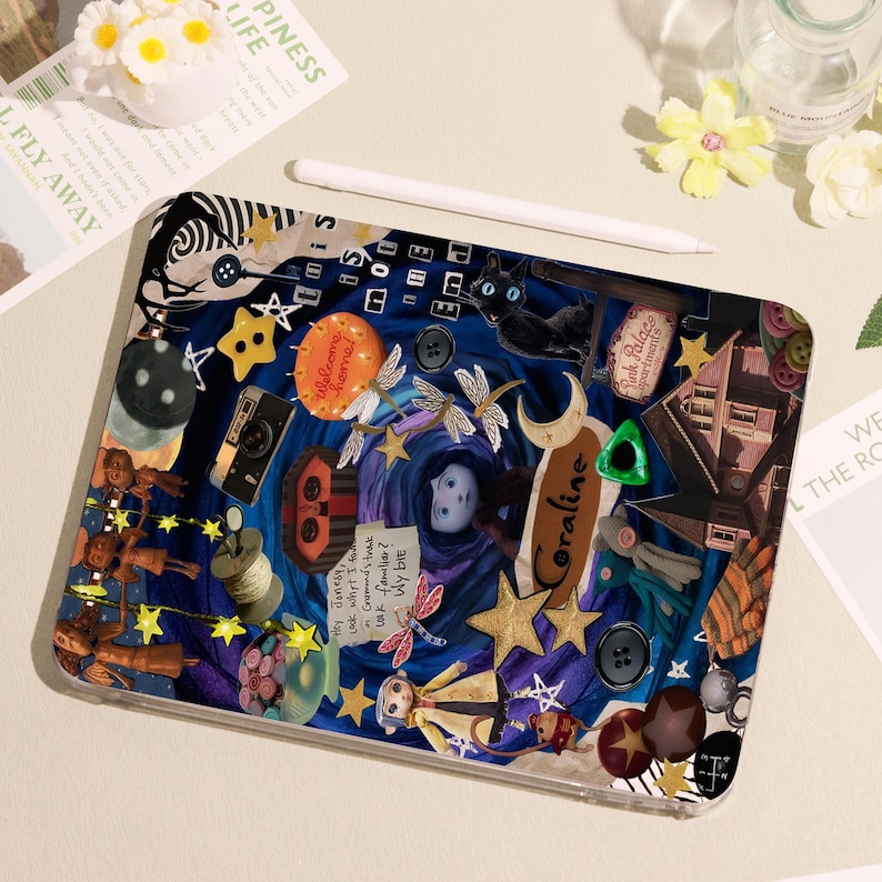 May include: A decorative tablet case featuring a collage design with a dark blue background. The design includes stars, buttons, a camera, and the word "Coraline". A white stylus rests on top of the case.