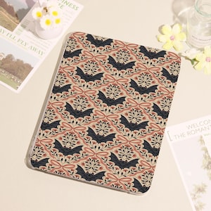May include: A tablet case featuring a repeating pattern of black bats, pink bows, and floral elements on a beige background. The design is symmetrical and covers the entire surface of the case.