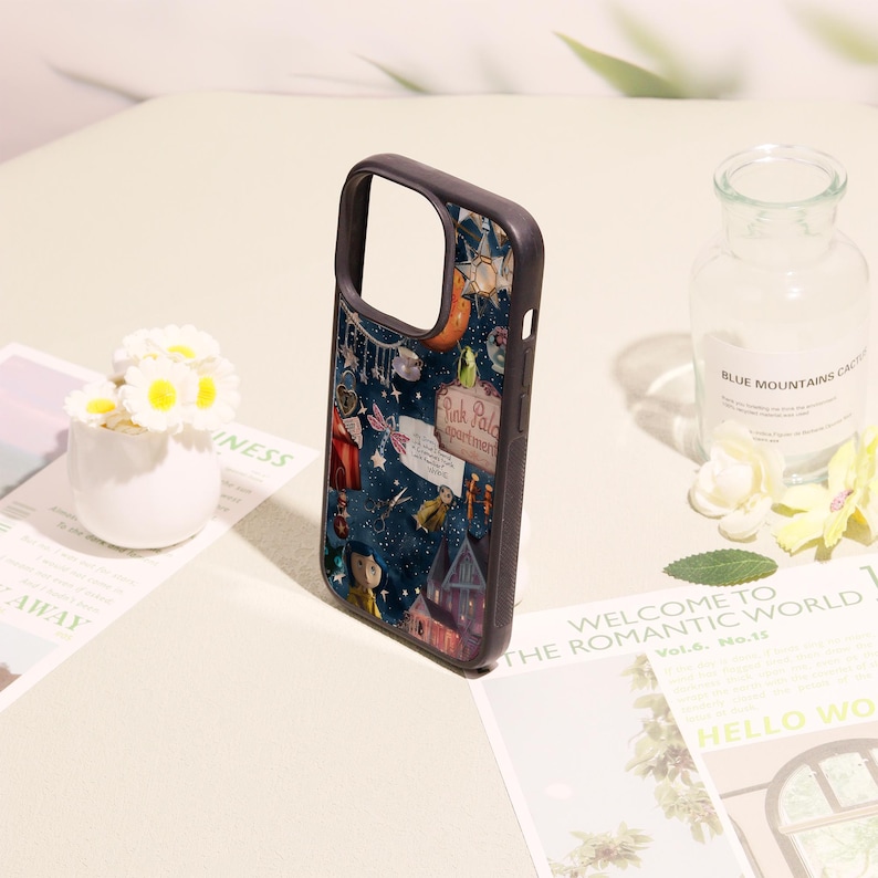 May include: A dark-themed phone case featuring a collage of illustrations including a character in a yellow raincoat, a house, a sign reading Pink Palace apartments, scissors, and various celestial elements against a deep blue starry background.