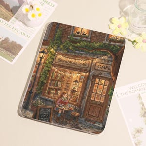 May include: A tablet case with an illustration of a bookstore cafe. The artwork shows a person reading at a table outside the cafe, a sign that says "Bookstore Cafe", and fairy lights. The colour palette includes warm browns and greens.