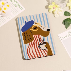May include: A tablet case featuring a cartoon dog wearing a blue beret and round glasses, holding a coffee cup. The dog is dressed in a red and white striped shirt, set against a background of blue and white vertical stripes.