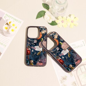 May include: Two dark-rimmed phone cases featuring a dark blue background with a whimsical collage of stars, a small house, a character with blue hair, scissors, and a sign that reads Pink Palace apartments, along with other small decorative elements.