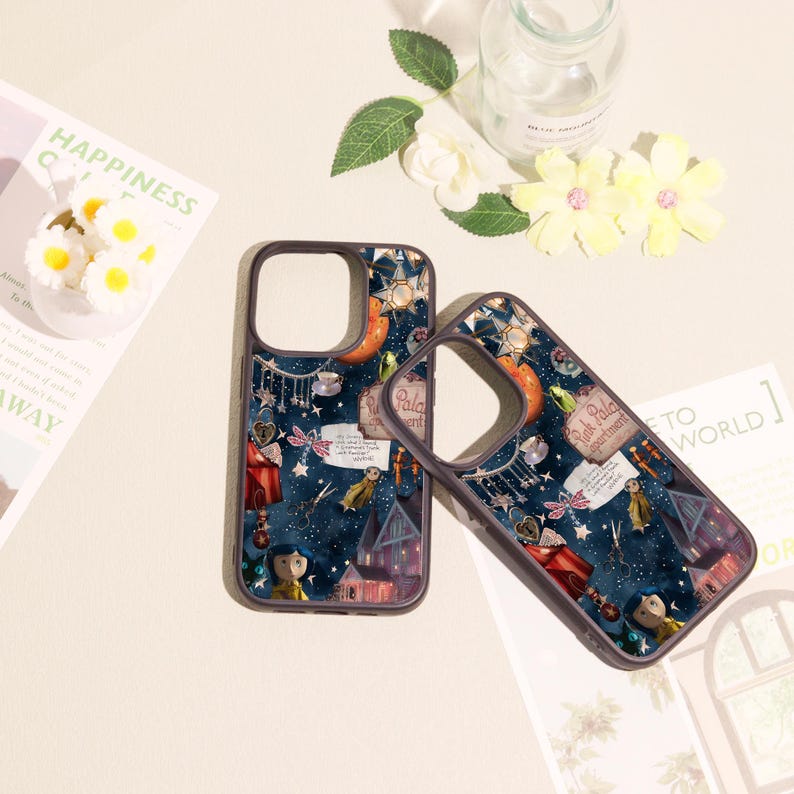 May include: Two dark-rimmed phone cases featuring a dark blue background with a whimsical collage of stars, a small house, a character with blue hair, scissors, and a sign that reads Pink Palace apartments, along with other small decorative elements.
