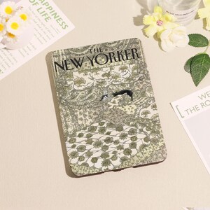 Puede incluir: Una funda rectangular con la portada de la revista "The New Yorker". El diseño presenta un estampado floral en tonos verdes y beige, con el título de la revista. Un gato negro también es visible en el diseño.