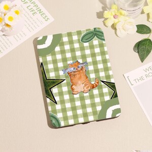 May include: A green and white checkered tablet case featuring a cartoon orange tabby cat holding two fish. The case has green star and leaf accents. The text "APPINESS OF LIFE" is visible on a nearby paper.