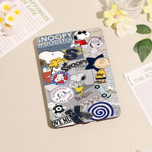 Op de afbeelding: Een tablethoesje met een collage-ontwerp met Snoopy en Peanuts-personages. Het ontwerp bevat Snoopy, Woodstock, Charlie Brown en andere personages, samen met het Converse All Star-logo. De hoes heeft een blauw en wit kleurenschema.