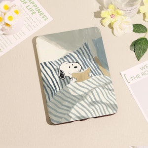 May include: A rectangular case featuring a cartoon Snoopy character reading a book in bed. The case has a blue and white striped pattern, with a light blue and beige background. The image also includes text from a magazine.