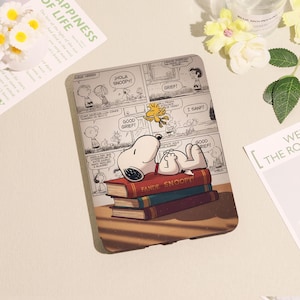 May include: A tablet case with a Snoopy comic strip design. Snoopy is lying on books with "FANDE SNOOPY" visible. The case has a light brown border and a comic strip design with speech bubbles. The case is likely for a tablet.