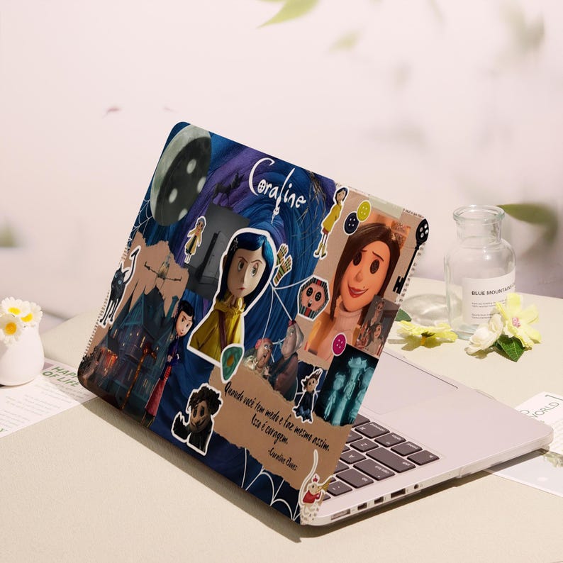 May include: A laptop with a Coraline-themed cover featuring character illustrations, a moon, and the word "Coraline." The cover has a collage design with a quote in Portuguese. The laptop is on a white surface.