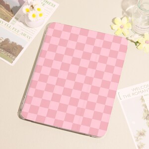 May include: A pink and light pink checkered tablet case. The case is rectangular with rounded corners and a clear border. The case is on a light beige surface, with a small vase of white flowers and a magazine.