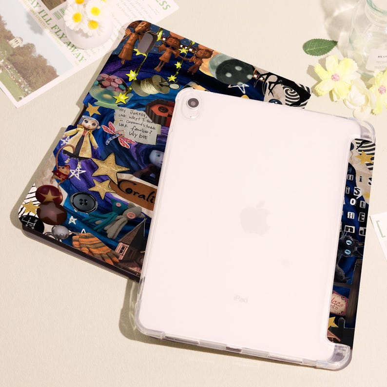 May include: A white iPad with a clear protective case rests on top of a patterned case. The patterned case features a collage of illustrations, including stars, dolls, and text. The text includes the words "Coralie" and "Wy bre."
