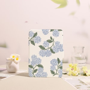 May include: A rectangular, off-white notebook or journal with a floral pattern of blue hydrangea blooms and green leaves. The notebook is the focal point, with a soft, neutral background.