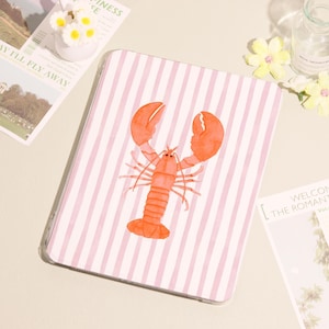 May include: A notebook featuring a pink and white striped design with a vibrant orange lobster illustration. The lobster is centrally positioned. Other decorative items are visible around the notebook.