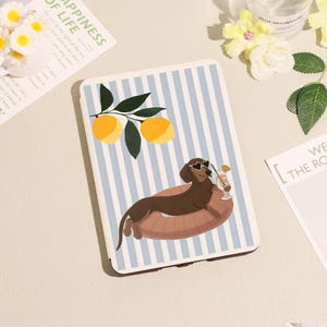 Könnte beinhalten: Eine Tablet-Hülle mit blau-weißem Streifenmuster, die einen Cartoon-Dackel mit herzförmiger Sonnenbrille und einem Cocktail zeigt. Die Hülle hat auch Illustrationen von Zitronen und Blättern.