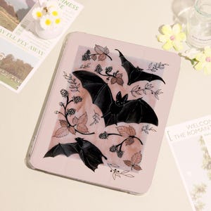 May include: A light pink tablet case featuring a watercolor design of black bats, leaves, and berries. The case has a clear border and a soft, muted color palette. The design is repeated across the surface.