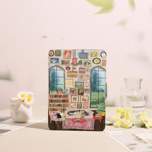 May include: A tablet case with a detailed illustration of a room interior. The design includes a bookshelf, artwork, windows, and a sofa with cats and a dog. The colour palette is warm, with a variety of framed pictures.