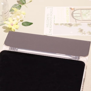 May include: A clear tablet case with a gray cover and a black screen. A white stylus is visible inside the case. The text "WELCOME TO THE ROMANTIC WORLD" is on a card in the background. Yellow flowers and a bottle are also in the background.