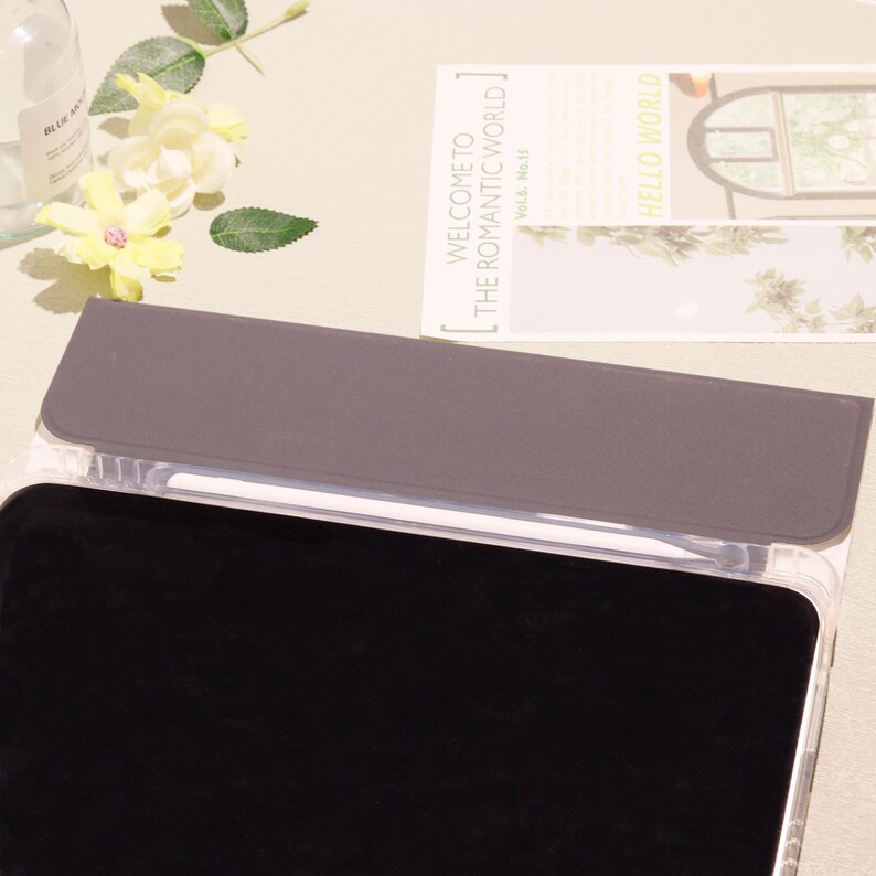 May include: A clear tablet case with a gray cover and a black screen. A white stylus is visible inside the case. The text "WELCOME TO THE ROMANTIC WORLD" is on a card in the background. Yellow flowers and a bottle are also in the background.