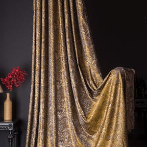 May include: Luxurious gold and brown patterned curtain fabric. The fabric is draped, revealing a detailed, swirling design. The curtain hangs in front of a dark wall, with a small table and vase of red flowers visible.