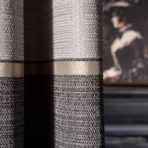 May include: Close-up of textured gray and brown curtains with a gold band. The curtains have a woven pattern and are partially closed, revealing a glimpse of a framed portrait in the background. The fabric appears thick and durable.