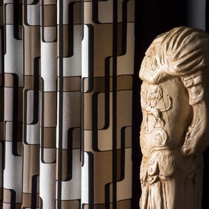Könnte beinhalten: Eine beige Statue einer klassischen Figur steht neben einem gemusterten Vorhang. Der Vorhang hat ein sich wiederholendes geometrisches Design in Braun-, Schwarz- und Weißtönen. Die Statue hat detaillierte Schnitzereien und ist seitlich beleuchtet.