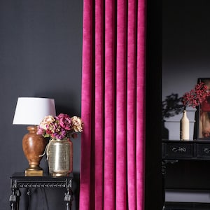 Könnte beinhalten: Ein lebendiger magenta Vorhang mit vertikalen Falten. Ein dunkler, verzierter Beistelltisch trägt eine Lampe mit weißem Schirm, eine goldene Vase mit Blumen und eine dunkle Wand im Hintergrund.