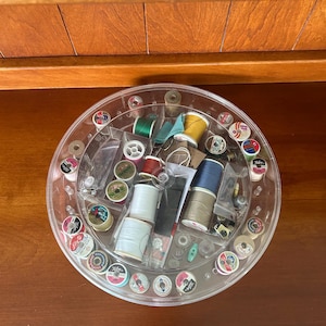May include: A circular, clear plastic rotating sewing organizer filled with various spools of thread in colors including white, blue, yellow, green, and red, along with small sewing notions like buttons and needles stored in divided compartments.