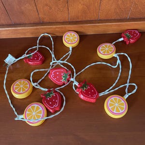 Puede incluir: Una cadena de luces decorativas con fresas rojas y rodajas de cítricos amarillos alternadas. Las luces están conectadas por un cable blanco y tienen un enchufe estándar.