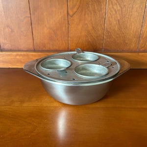 May include: A stainless steel egg poaching pan featuring a rounded base and a removable insert with three circular cups. The insert has small ventilation holes around the perimeter, and the main pan includes two side handles for lifting.