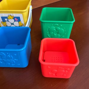 May include: Four brightly coloured plastic stacking cups. The cups are yellow, green, blue, and red. The yellow cup has a white handle and a sticker with cartoon chicks. The green, blue, and red cups have embossed animal designs.