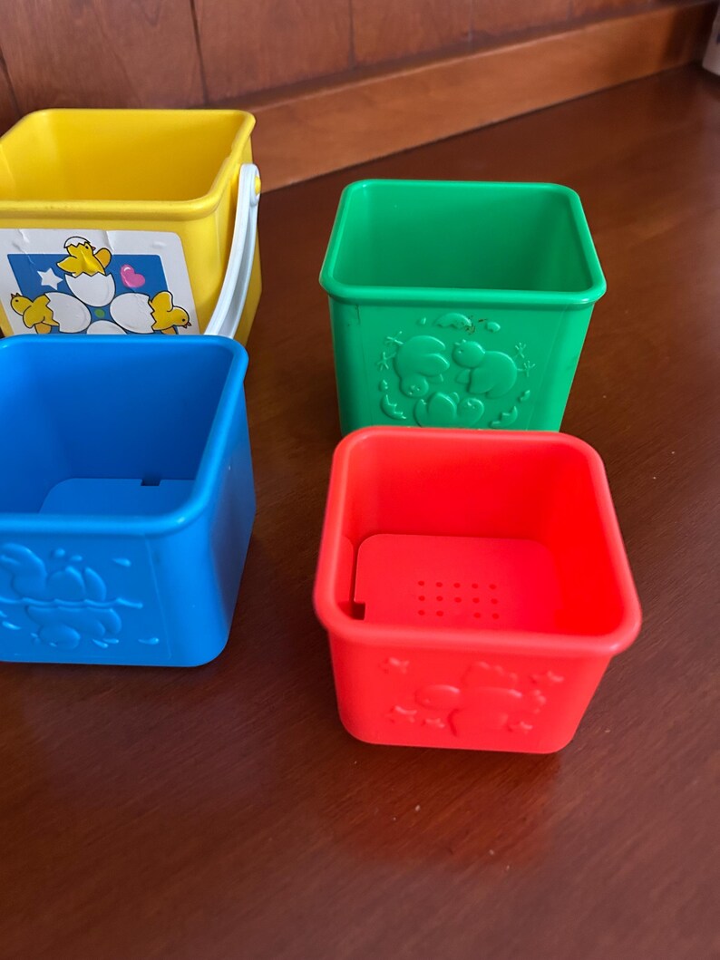 May include: Four brightly coloured plastic stacking cups. The cups are yellow, green, blue, and red. The yellow cup has a white handle and a sticker with cartoon chicks. The green, blue, and red cups have embossed animal designs.