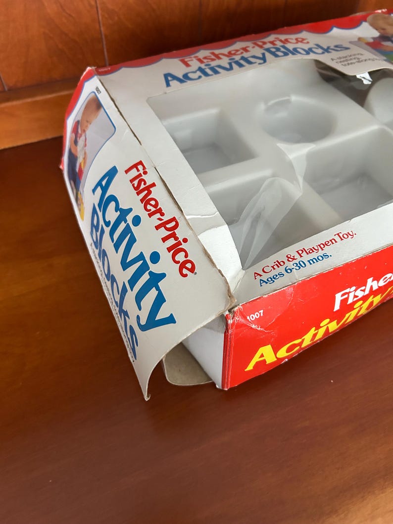 May include: A vintage Fisher-Price Activity Blocks toy box. The box is red, white, and blue with the words "Fisher-Price Activity Blocks" in bold lettering. The box is slightly open, revealing the plastic toy blocks inside. The box is designed for ages 6-30 months.