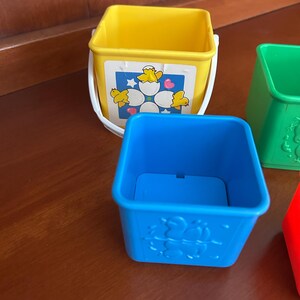 May include: A set of colourful, stackable toy buckets. The yellow bucket has a white handle and a graphic of chicks hatching from eggs. The blue bucket has a moulded design of chicks. Other buckets are green and red.