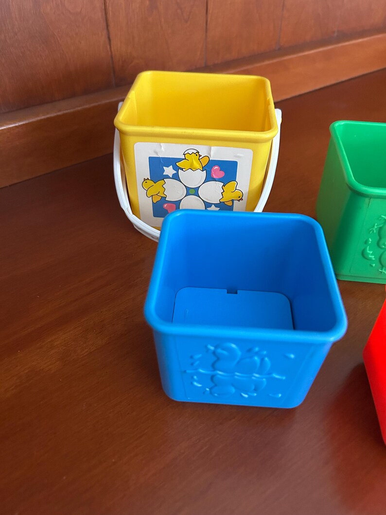 May include: A set of colourful, stackable toy buckets. The yellow bucket has a white handle and a graphic of chicks hatching from eggs. The blue bucket has a moulded design of chicks. Other buckets are green and red.