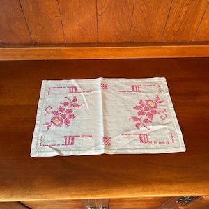 Può includere: Una tovaglietta rettangolare in lino color bianco sporco con un motivo a punto croce nei toni del rosa e del giallo. Il design presenta due motivi floreali sui lati sinistro e destro, completati da accenti geometrici lungo i bordi.
