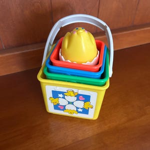 May include: A colourful stacking toy featuring a yellow bucket with a white handle, and green, blue, and red containers. The top piece is a yellow chick in a white eggshell. The bucket has a chick and egg graphic.