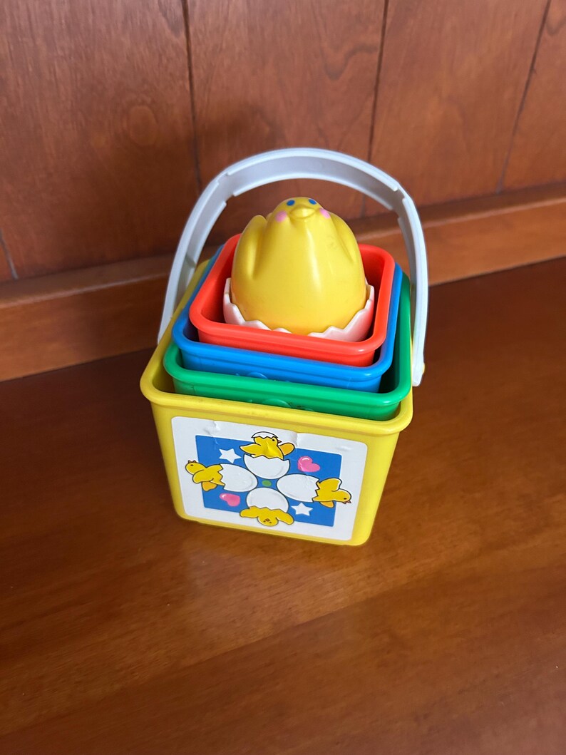 May include: A colourful stacking toy featuring a yellow bucket with a white handle, and green, blue, and red containers. The top piece is a yellow chick in a white eggshell. The bucket has a chick and egg graphic.