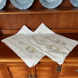 May include: Two white linen napkins with embroidered yellow floral designs. The napkins are folded and displayed on a wooden cabinet. Blue and white patterned plates are visible in the background.