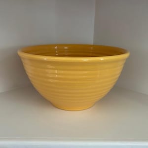 May include: A bright yellow ceramic bowl with a ribbed exterior. The bowl is round and sits on a white shelf. The bowl is empty and appears to be new. The bowl is a kitchenware item.