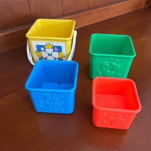 May include: A set of four brightly coloured plastic stacking buckets. The buckets are yellow, green, blue, and red. The yellow bucket has a white handle and a sticker with a flower and chick design. The other buckets have embossed animal designs.