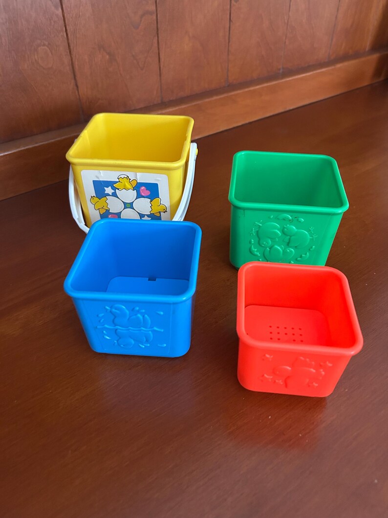 May include: A set of four brightly coloured plastic stacking buckets. The buckets are yellow, green, blue, and red. The yellow bucket has a white handle and a sticker with a flower and chick design. The other buckets have embossed animal designs.