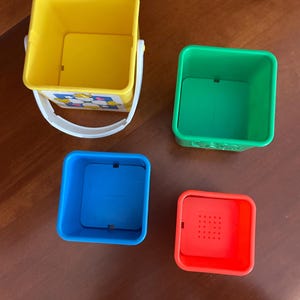May include: Four brightly coloured stacking toy blocks. The largest is yellow with a white handle, the next is green, followed by blue, and the smallest is red with holes. The toys are square and designed for children.